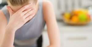 a woman with a migraine stands at her kitchen counter and holds her head in pain