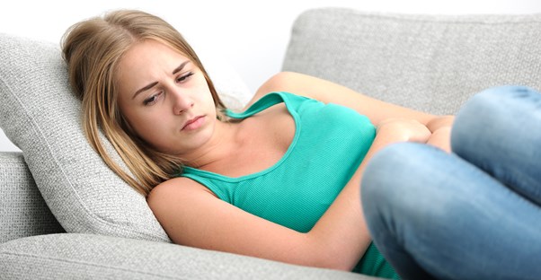 a young woman lying on a couch holds her stomach in pain from crohn's disease or ulcerative colitis